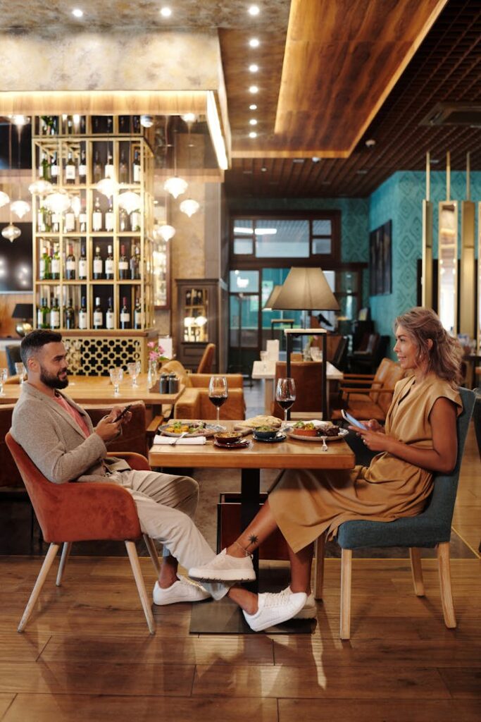 Couple enjoying a meal and conversation at a stylish restaurant, using smartphones and wine glasses present.